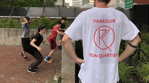 Un homme de dos avec la mention « Parkour ton quartier » sur son chandail. Devant lui, plusieurs enfants se préparent è sauter par-dessus un mur en béton.