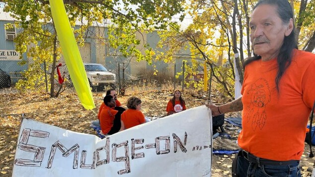 Un homme avec un chandail orange qui tient un affiche qui dit : Smudge On 