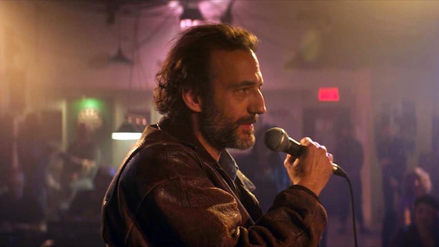 Un homme (Martin Dubreuil) portant une veste en cuir et tenant un micro, sur une scène, de profil.