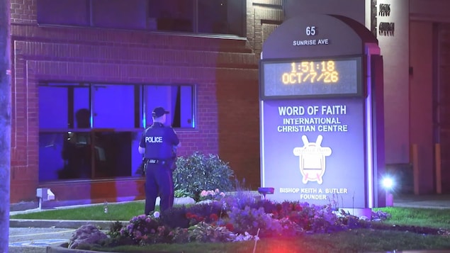 Un policier se tient devant un centre religieux de Toronto, au 65, avenue Sunrise. La façade est éclairée par les gyrophares bleus et rouges des véhicules d’urgence.