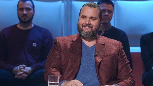 Un homme qui sourit. Il porte un chandail bleu et un veston brun à fines lignes bleues.