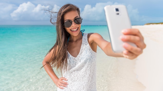 Une jeune femme portant des lunettes soleil prend un autoportrait avec son téléphone, devant une plage et la mer bleue des Caraïbes.