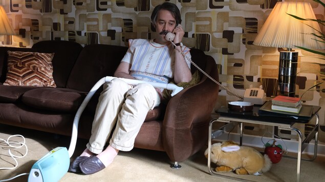 Un homme portant un tablier assis dans un canapé parle au téléphone.