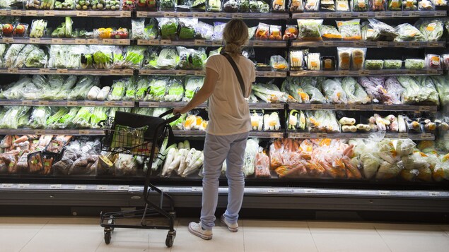 Une cliente poussant un panier regarde des légumes emballés dans des pellicules plastique dans une épicerie.