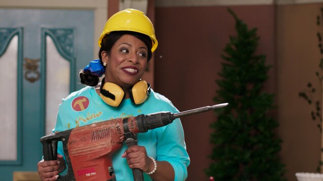 Elle tient un grosse perceuse et porte un casque de la construction sur la tête. 