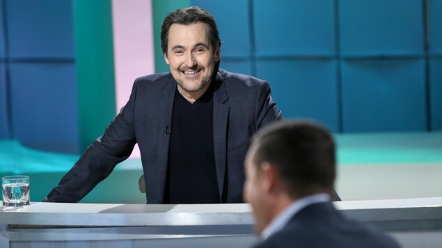 Il sourit à Dany Turcotte. Il porte un chandail et un veston sombres. Il sourit à Dany Turcotte. Il porte un chandail et un veston sombres.