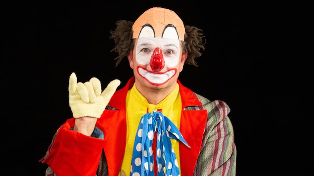 Un clown avec un nez rouge, une chemise jaune, une cravate bleue à pois blancs, des pantalons rouges et un veston à rayures colorées.