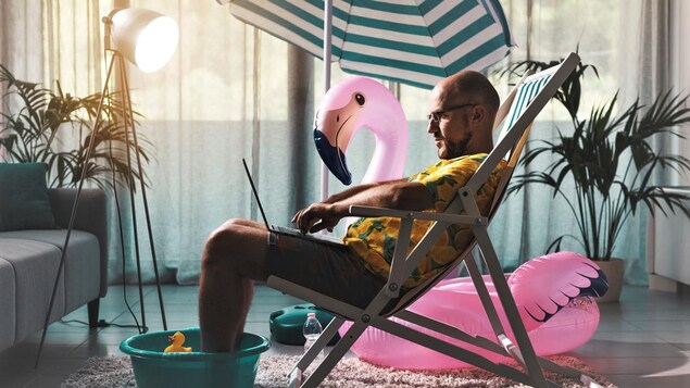 Un homme assis dans une chaise de plage à l'intérieur de sa maison. Les pieds de l'homme trempent dans un seau d'eau. Un immense flamant rose gonflable est à ses côtés.