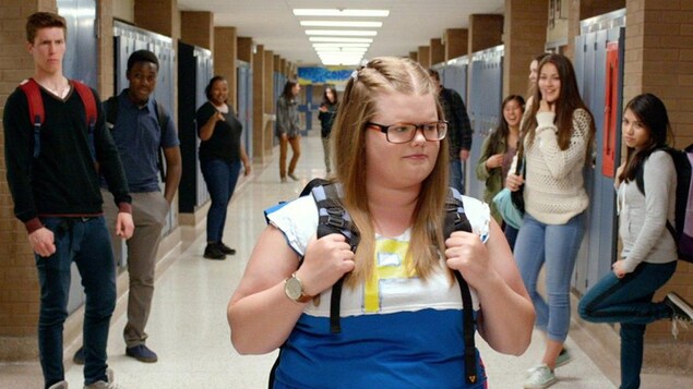 Une jeune fille plus ronde marche au milieu d'un couloir dans un collège sous le regard moqueur d'autres élèves.