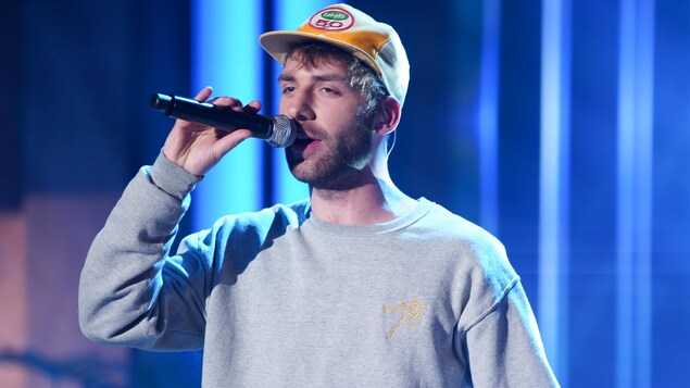 Un homme, qui porte une casquette Labatt 50. Il chante en tenant un micro dans sa main.