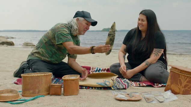 Sur une plage, un aîné montre des artéfacts à une jeune femme.