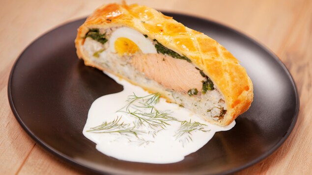 Un koulibiac servi sur une assiette ronde accompagné d'une sauce blanche.