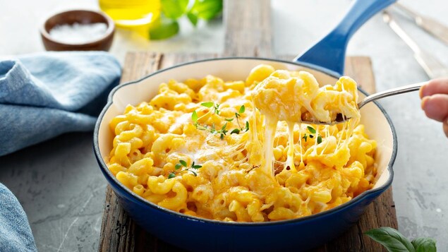 Un macaroni au fromage dans une poêle bleue. Une main tient une fourchette qui contient du macaroni.