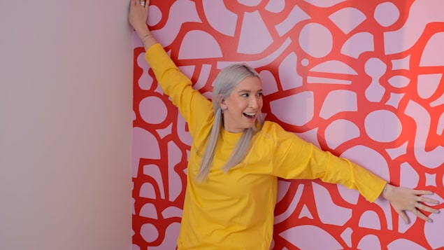 Marianne Plaisance se trouve devant un mur avec du papier peint et elle ouvre les bras. 