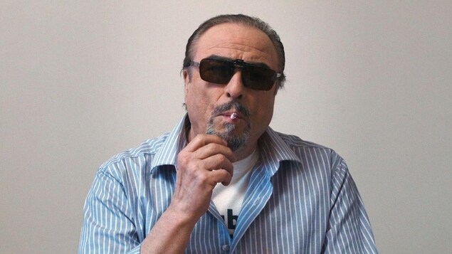 Un homme (Dino Tavarone) portant des lunettes de soleil et fumant une cigarette, en plan de face.