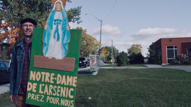 Un homme tient une affiche où l'on peut lire : Notre-Dame-de-l'Arsenic priez pour nous­.
