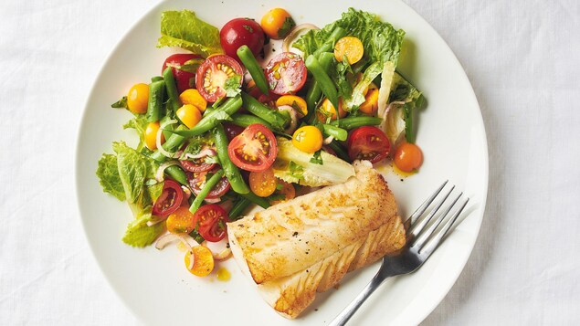 Dans une assiette on voit un morceau de poisson blanc roti et une salade colorée de tomates cerise, fèves et cerises de terre.