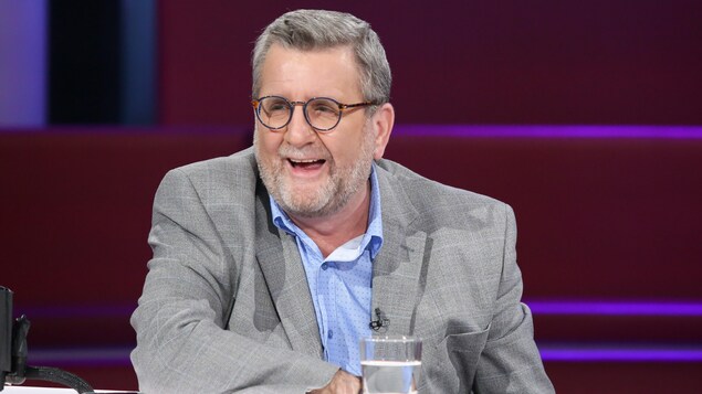 Un homme qui rit. Il porte une chemise bleue et un veston gris.