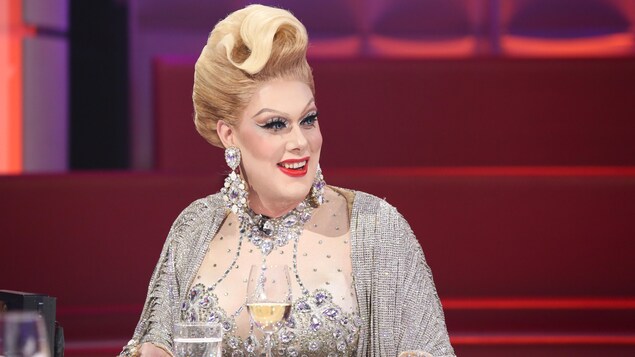 Une drag queen qui porte une perruque blonde et une robe parsemée de pierres brillantes.