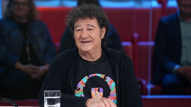 Un homme aux cheveux frisés. Il porte un t-shirt et un chandail à capuchon.