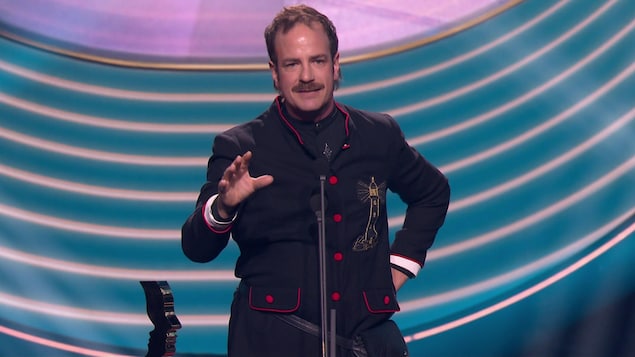Robin-Joël Cool sur scène. Il parle dans un micro et lad   prix Gémeaux est à côté de lui.