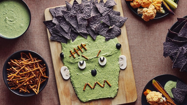 Une trempette en forme de visage de Frankenstein sur une planche de bois est accompagnée d’un bol de trempette, d’un bol de pretzels, d’un bol de croustilles de maïs bleu et de deux assiettes de légumes.