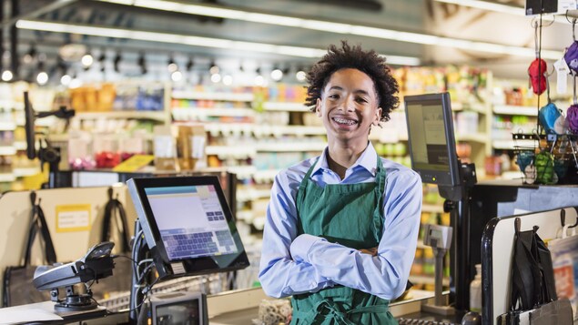 Un employé adolescent debout devant sa caisse enregistreuse d'une épicerie. Il sourit. Il est afro-canadien.