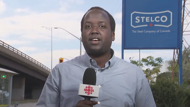 Un journaliste de Radio-Canada fait un reportage devant l’usine Stelco. Derrière lui, une grande enseigne bleue affiche le nom et le logo de l’entreprise.