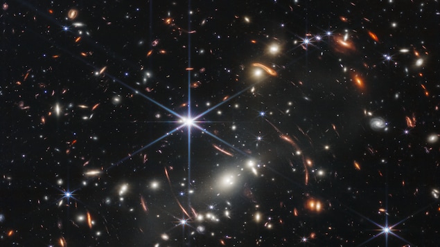 Le fond de l'espace est noir. Des milliers de galaxies apparaissent partout dans la vue. Leurs formes et leurs couleurs varient. Certains sont de différentes nuances d'orange, d'autres sont blancs. La plupart des étoiles apparaissent bleues et sont parfois aussi grandes que des galaxies plus éloignées qui apparaissent à côté d'elles. Une étoile très brillante est juste au-dessus et à gauche du centre.
