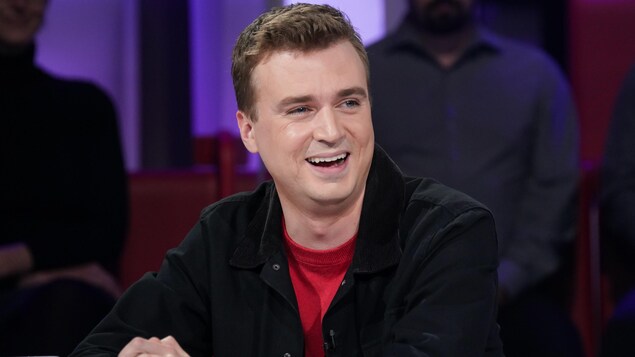 L'homme est en studio et répond aux questions de l'animateur. Il sourit.