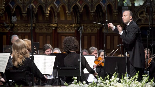 À droite, Yannick Nézet-Séguin se tient debout. Il tient dans sa main droite une baguette de chef d'orchestre. Des violonistes sont autour de lui, assis, en train de jouer. Les violonistes ont devant eux des lutrins avec des partitions de musique. En bas de l'image se trouvent des fleurs. Les musiciens se trouvent dans une église.