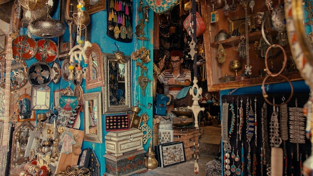 Un artisan est dans sa boutique, qui regorge de bijoux, de tableaux, de miroirs et de mains de Fatma. 