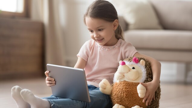 Elle a environ 5-6 ans et joue avec une tablette dans son salon, son nounours sous le bras.