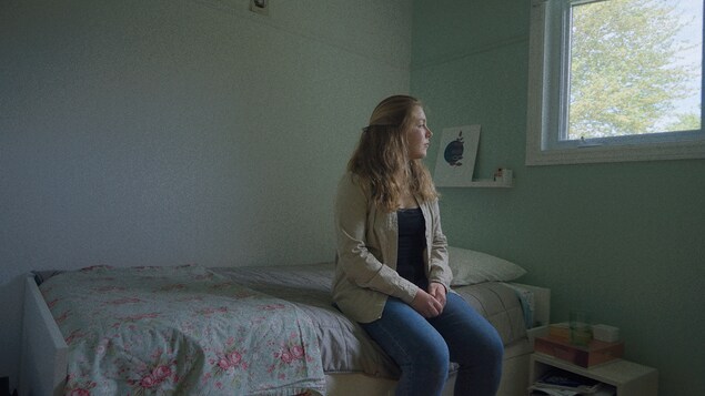 Une adolescente est assise sur son lit et regarde par le fenêtre.