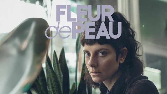 Devant une fenêtre et une plante verte, une femme détourne le regard vers la caméra.