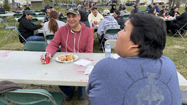 Une vingtaine de personnes mangent et jasent autour de tables communautaires à l'extérieur.