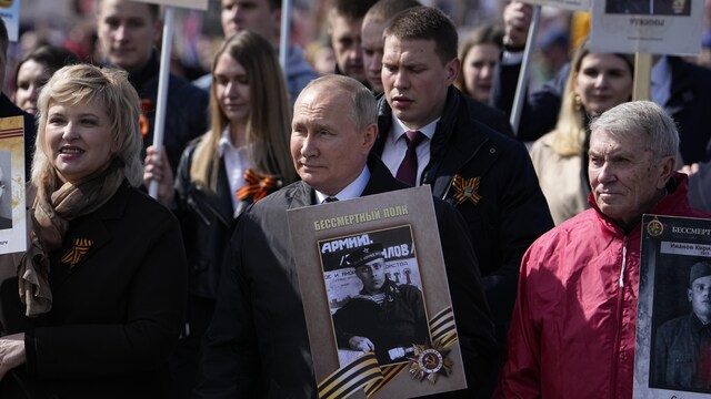 Le président russe Vladimir Poutine, entouré d'une foule de personnes portant des rubans rayés, tient une pancarte représentant un soldat à un défilé à Moscou.