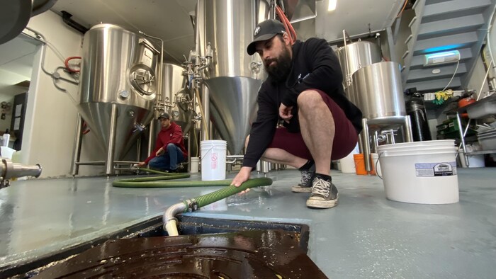 Accroupi, Martin Poirier tient le boyau qui transporte la levure brassicole de la cuve de brassage au drain.