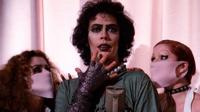Un homme androgyne est entouré de deux femmes portant des masques. 