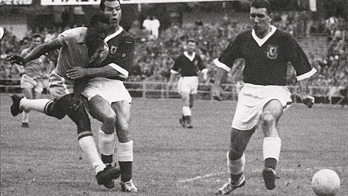 À gauche, Pelé lance le ballon près de deux joueurs de soccer adverses.