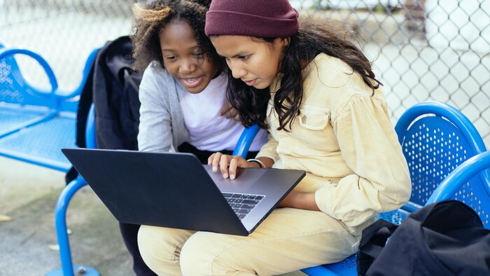 Deux jeunes filles assises sur un banc à l'extérieur avec un ordinateur portable sur les genoux.