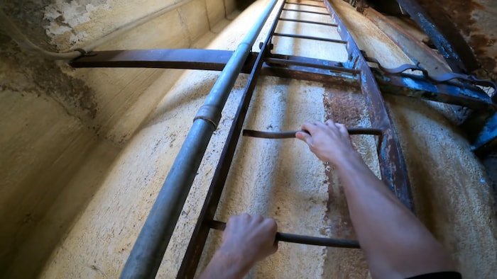 Les bras d'une personne agrippés à des barreau d'une échelle en métal située à l'intérieur d'une structure. 