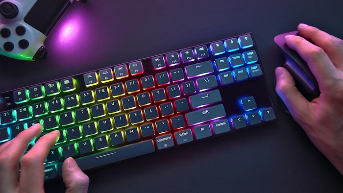 Des mains manipulant une souris, à côté d'un clavier illuminé en couleurs arc-en-ciel, à côté d'une manette de jeu vidéo.