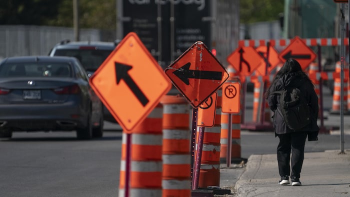 Une personne marche sur un trottoir près d'un chantier routier, à côté d'une série de cônes orange et de panneaux de signalisation.