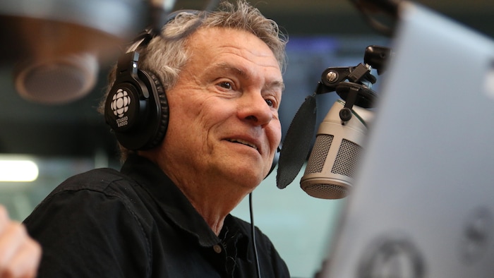 Paul Houde, casque d'écoute sur la tête, parle en souriant dans un studio de radio. 