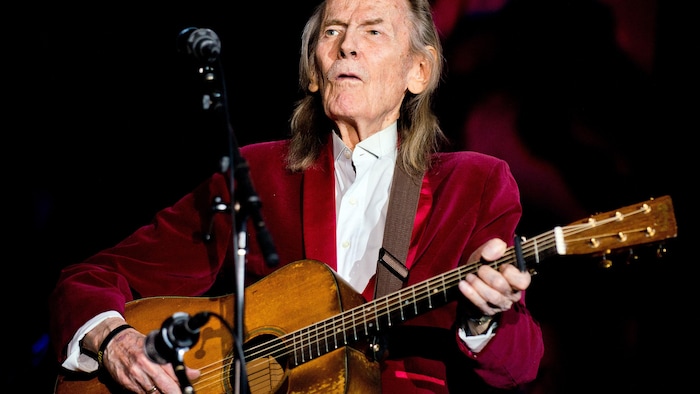 Gordon Lightfoot porte un veston rouge et joue de la guitare.