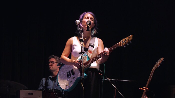 Gabrielle Papillon chante devant un micro et tient une guitare entre les mains.