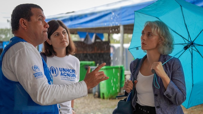 Un homme et une femme discutent avec une autre femme, qui tient un parapluie, à l'extérieur de tentes dans une station d'accueil de migrants.