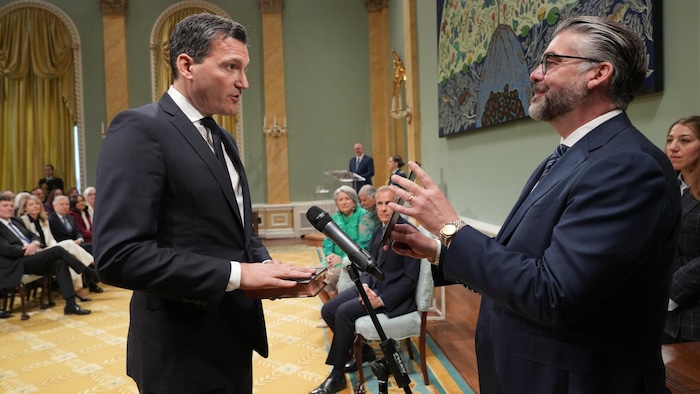 Dans une salle du Parlement, un homme nommé ministre prête serment en lisant un texte qu'un autre homme tient devant lui.