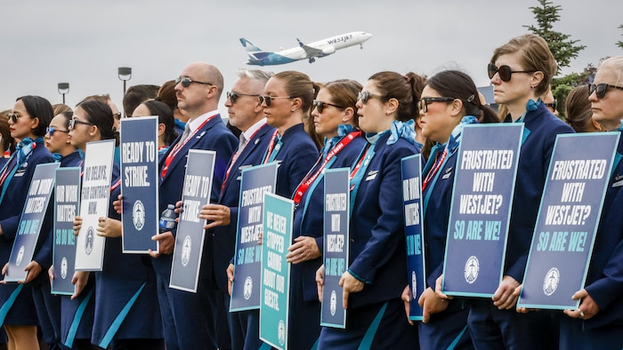 Une manifestation d’agents de bord de WestJet le 14 juillet 2026. On voit également un avion de l’entreprise décollé derrière eux.